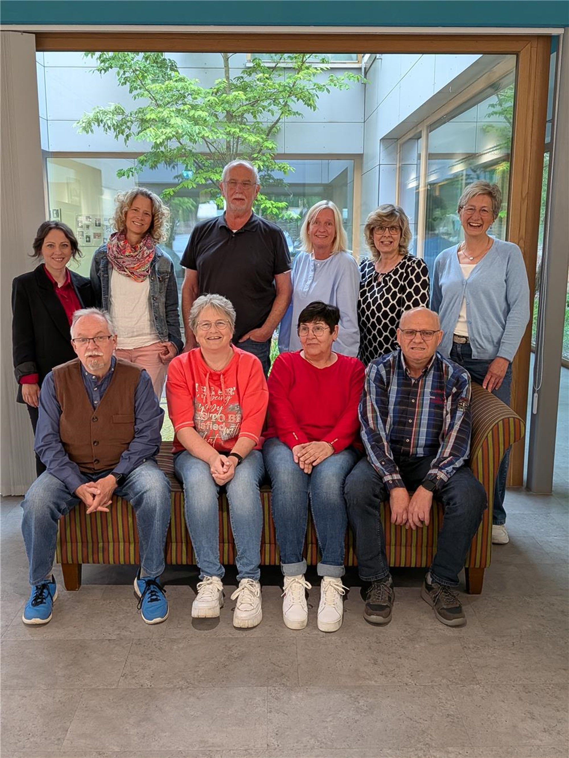 Das Team besteht aus drei Männern und sieben Frauen. Diese stehen vor einem großen bodentiefem Fenster. Ein Teil steht hinter einer Holzband, einige Teammitglieder sitzen darauf.