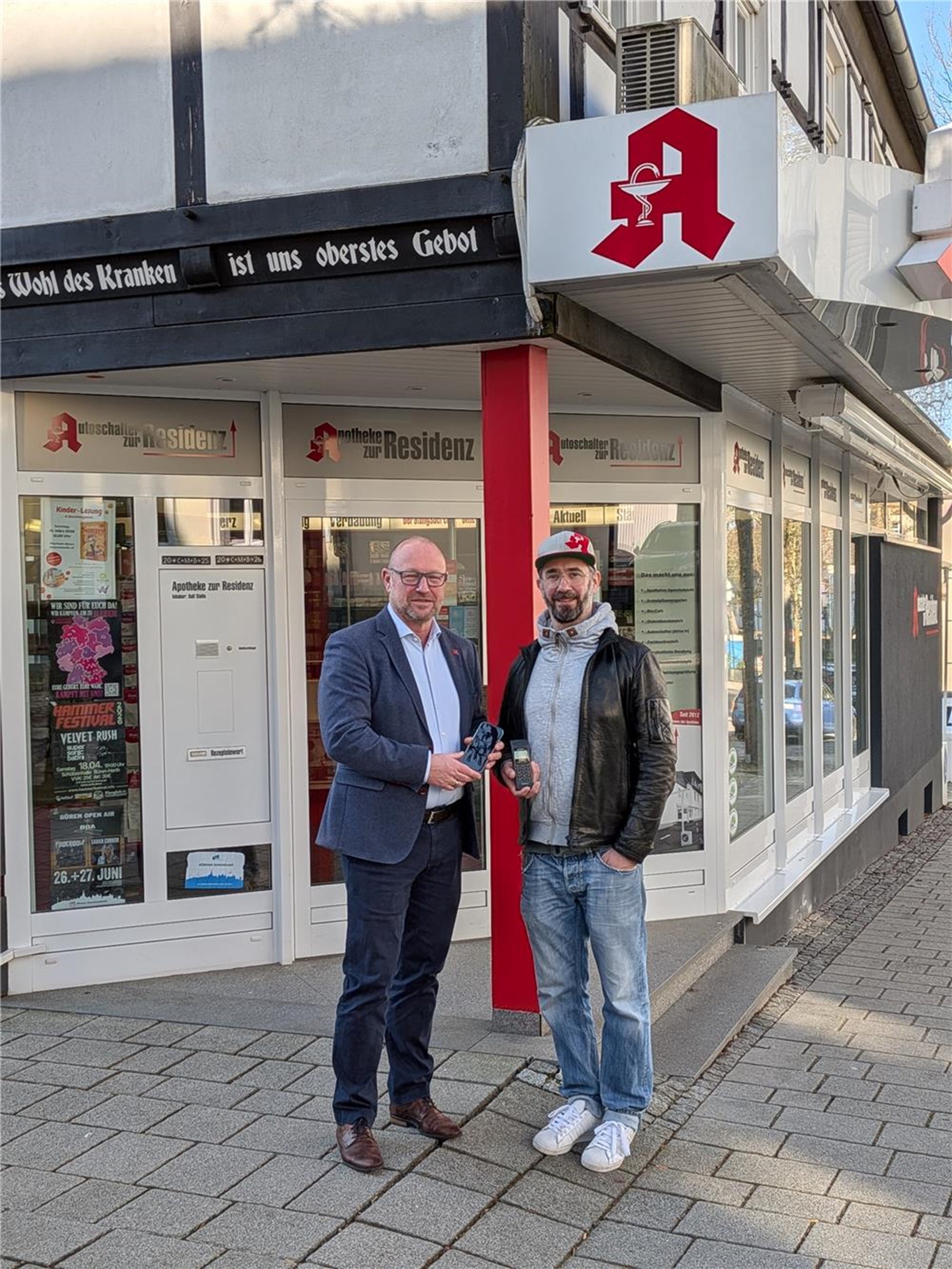 zwei Männer mittleren Alters, einer im Anzug, der andere leger mit basecap gekleidet stehen auf einem Treppenabsatz der Residenz-Apotheke und halten ihr Mobiltelefon als Hinweis auf die Digitalpaten