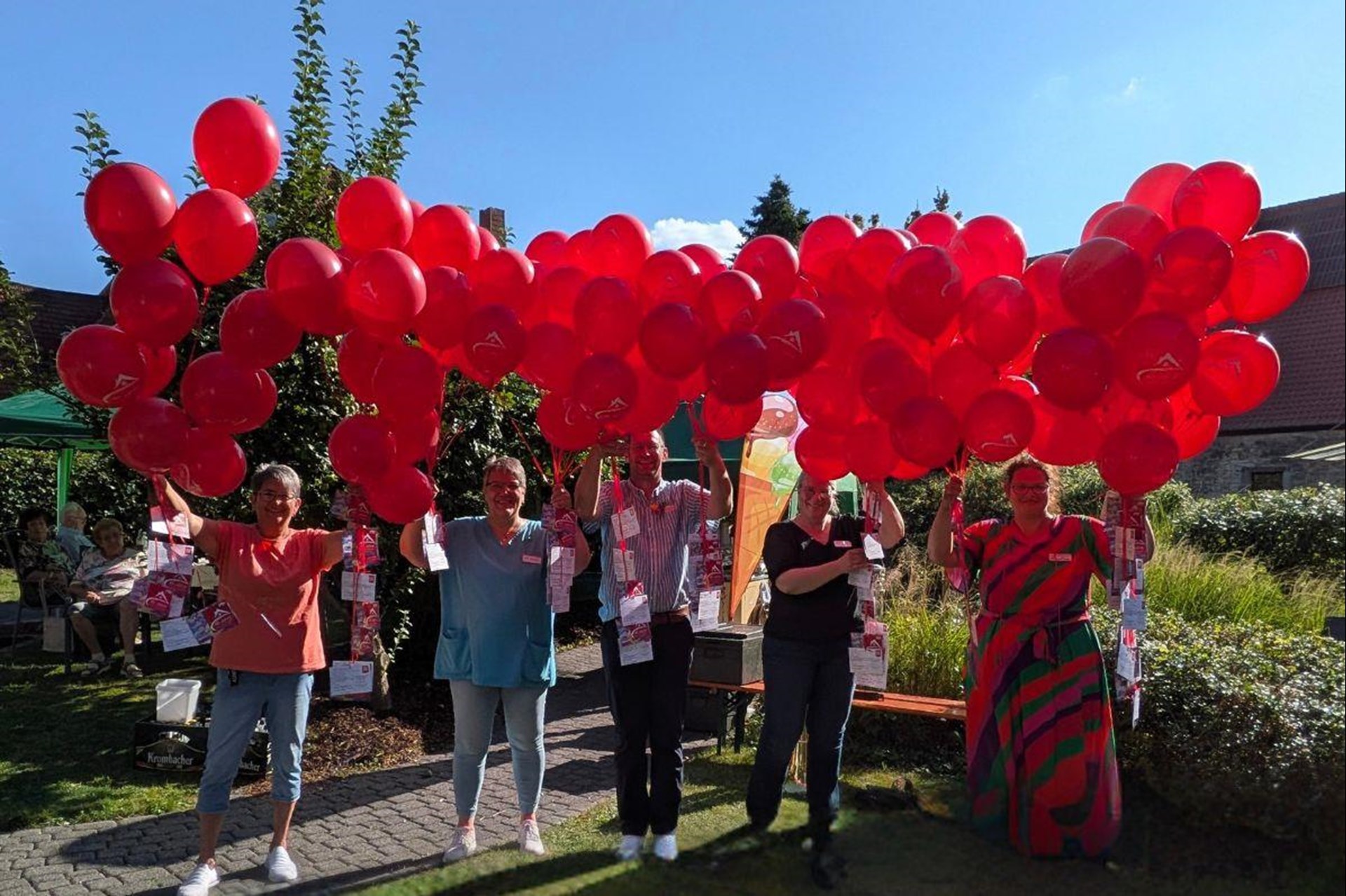 3 Frauen und 1 Mann stehen in einem Garten und halten jeder viele rote Luftballons in den ausgestreckten Händen. Die Ballons sind mit Wunschakarten versehen und sollen fliegen.