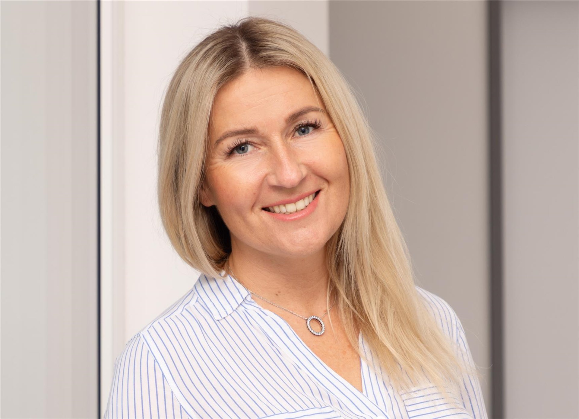 Frau mit langen blonden Haaren steht vor einem Fenster. Sie trägt ein weißes Oberteil