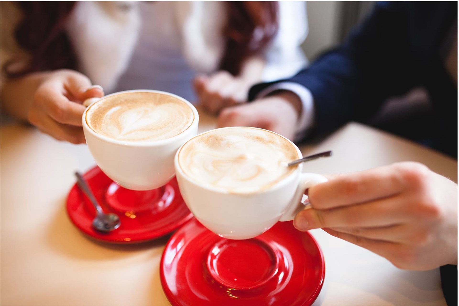 zwei rote Unterteller stehen auf einem hellen Tisch und zwei Tassen Kaffee mit Milchschaum werden hochgehalten. Zu sehen sind hier nur die Hände und Unterarme sowie die Tassen in der Hand.