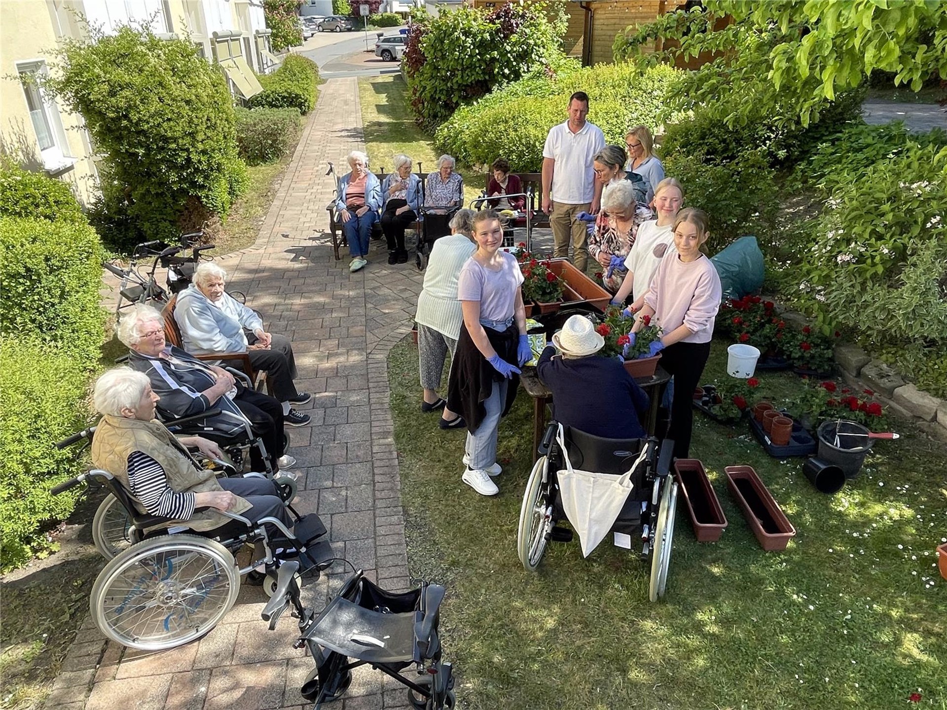 Seniorinnen und ehrenamtliche Schülerinnen sitzen und stehen an Tischen im Freien und bepflanzen Blumenkästen mit roten Geranien