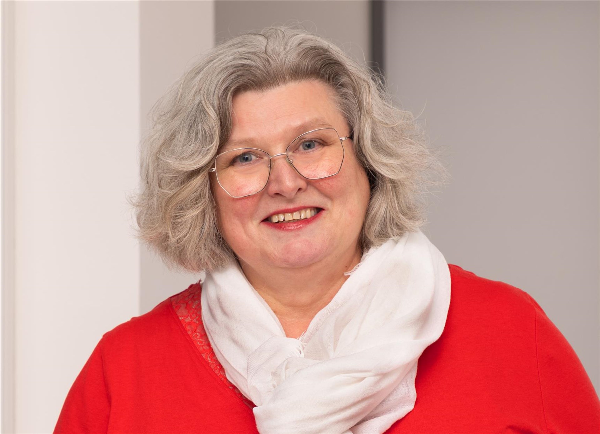 Frau mit schulterlangen grauenfarbenen Locken und Brille steht vor einem Fenster. Sie trägt ein weißes Oberteil und eine rote Jacke