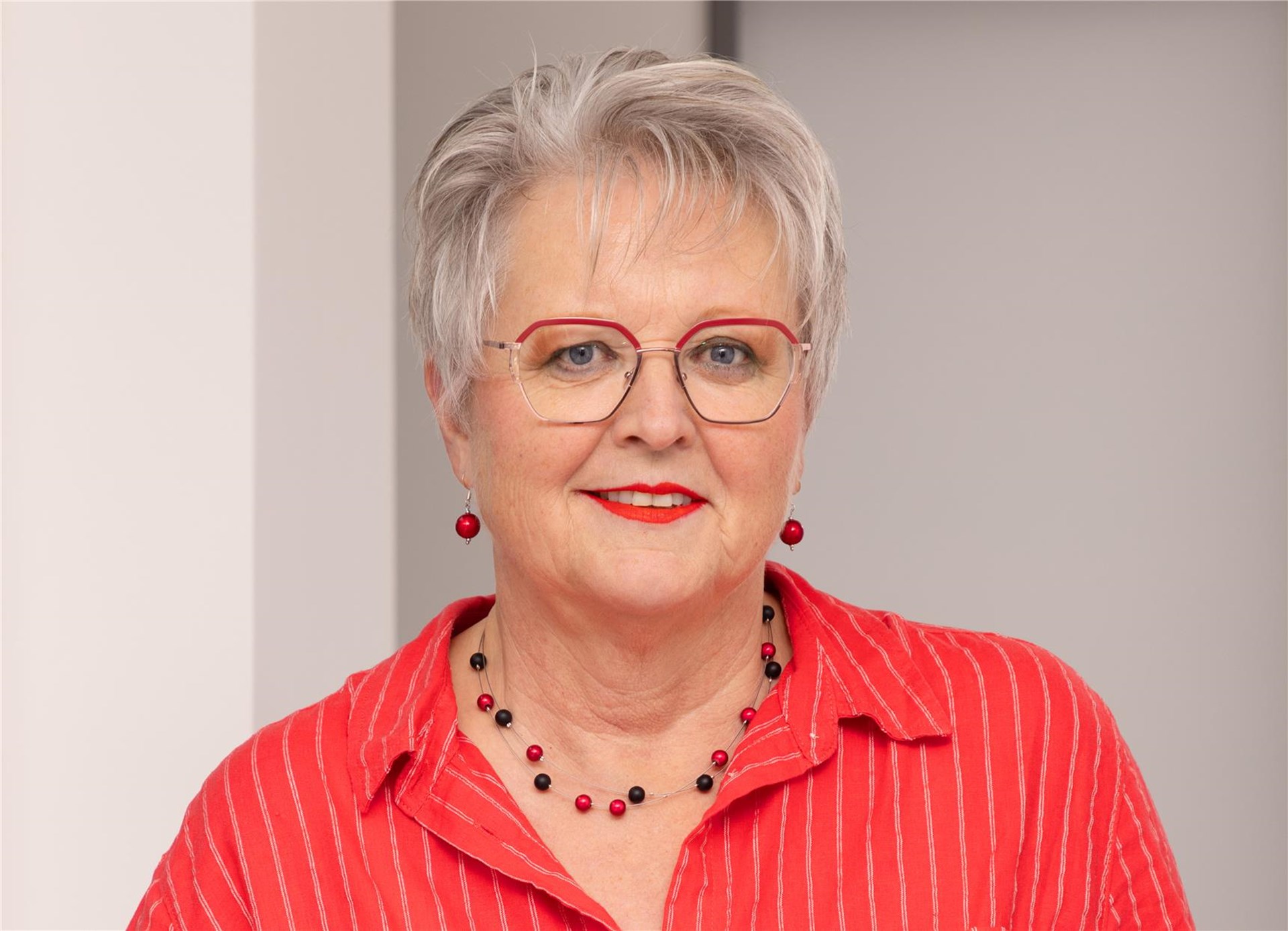 Frau mit kurzen blonden Jahren steht vor einem Fenster. Sie trägt ein weißes Oberteil mit roter Jacke