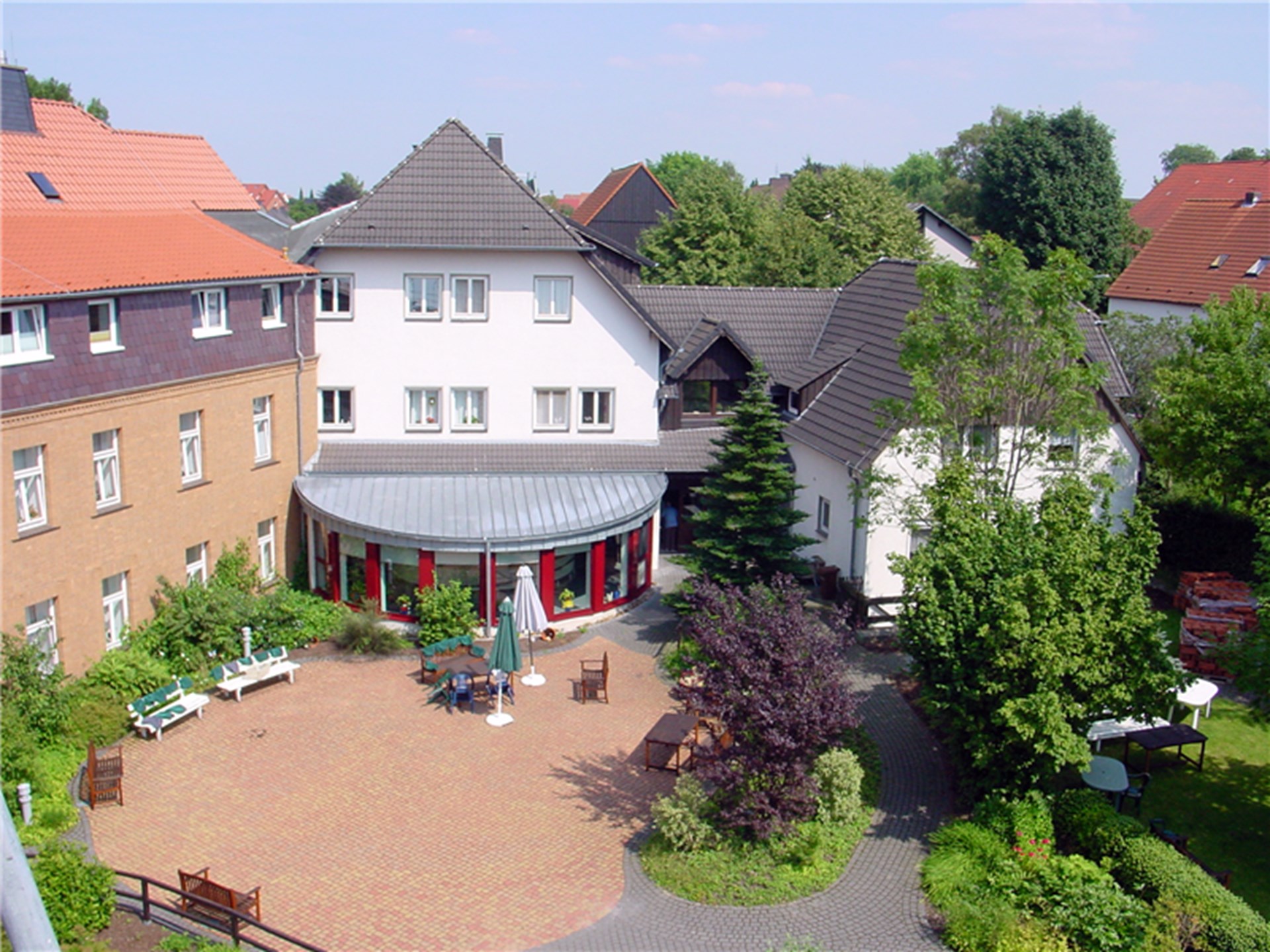 Luftaufnahme vom Innenhof des Altenheims "St. Clemens" Fürstenberg. Die weiße Aussenfassade wird durch ein rot akzentuiertes verglastes Rondell erweitert. Davor liegt ein grau gepflasterter Hof mit Grünanlagen