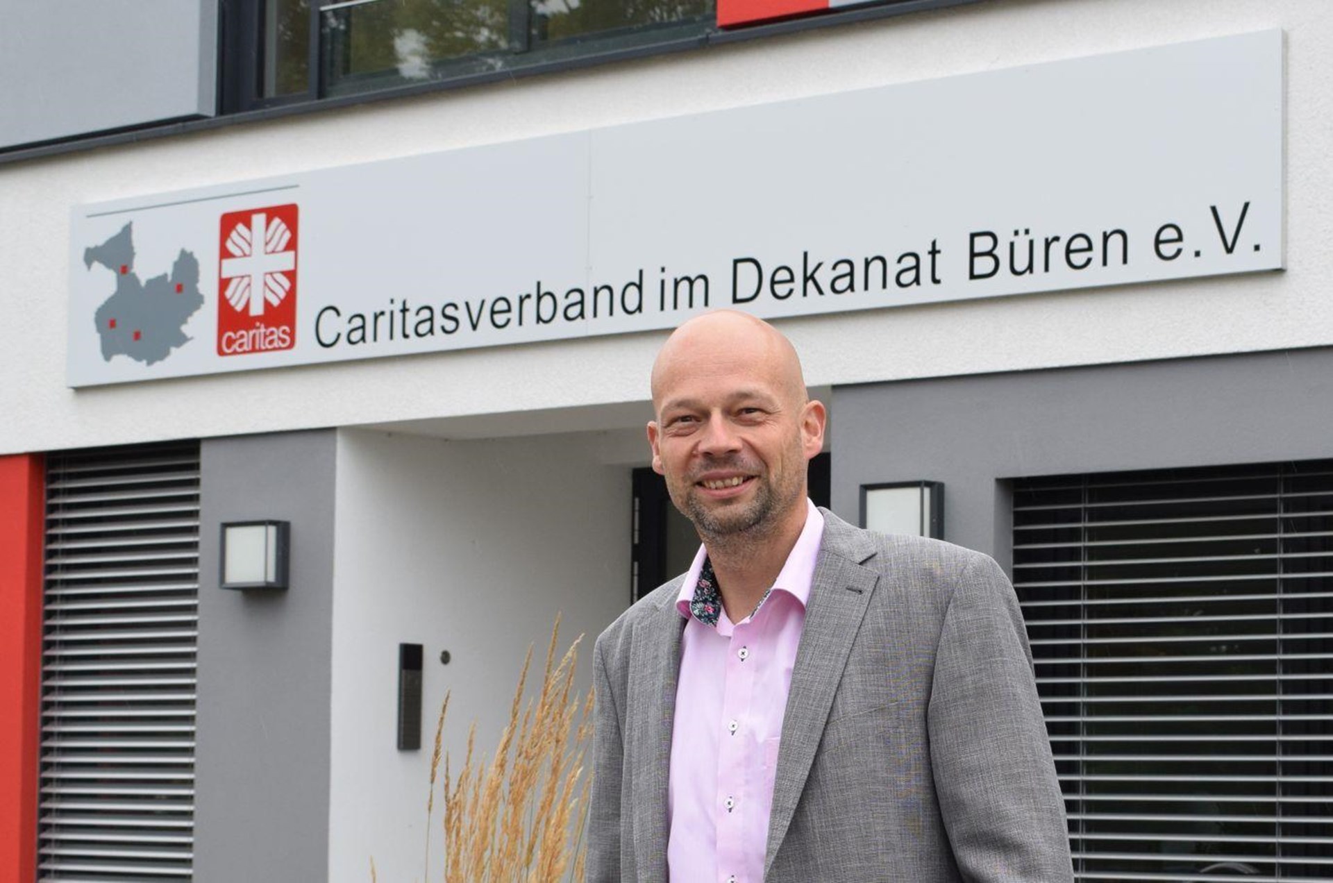 schlanker Mann mit Glatze, grauem Blazer, rosfarbenen Hemd steht vor dem Caritasgebäude