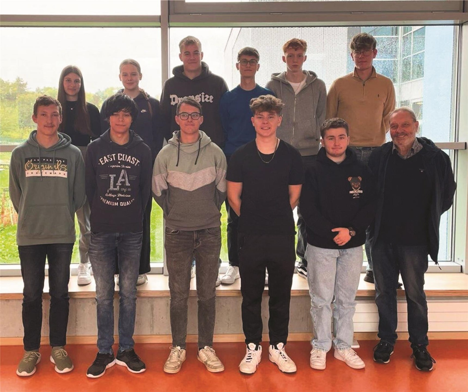 11 Schülerinnen und Schüler der Q1 sowie ein Lehrer der Gesamtschule Büren stehen vor einer Glaswand in der Schule