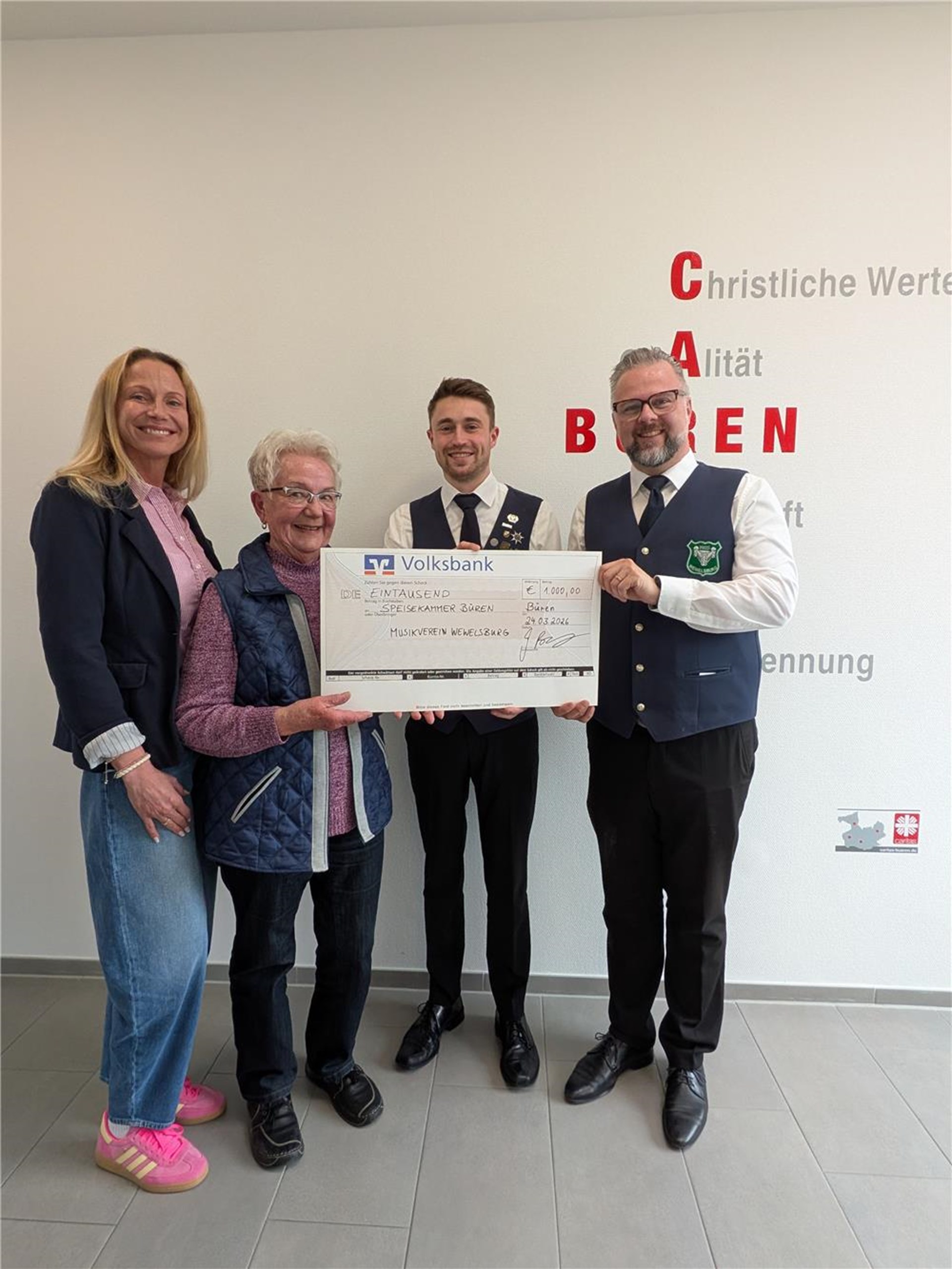 zwei Frauen und zwei Männer stehen vor einer weißen Wand mit Caritas Zeichen in rot. eine Frau, groß, blond mit welligem Haar, die andere klein, kurze graue Haare. Die beiden Herren tragen die Tracht des Musikvereins Edelweiß...dunkle Hosen, dunkle Westen, weiße Hemden. Zusammen halten Sie einen obligatorischen Scheck mit einem Wert von 1000 Euro in den Händen