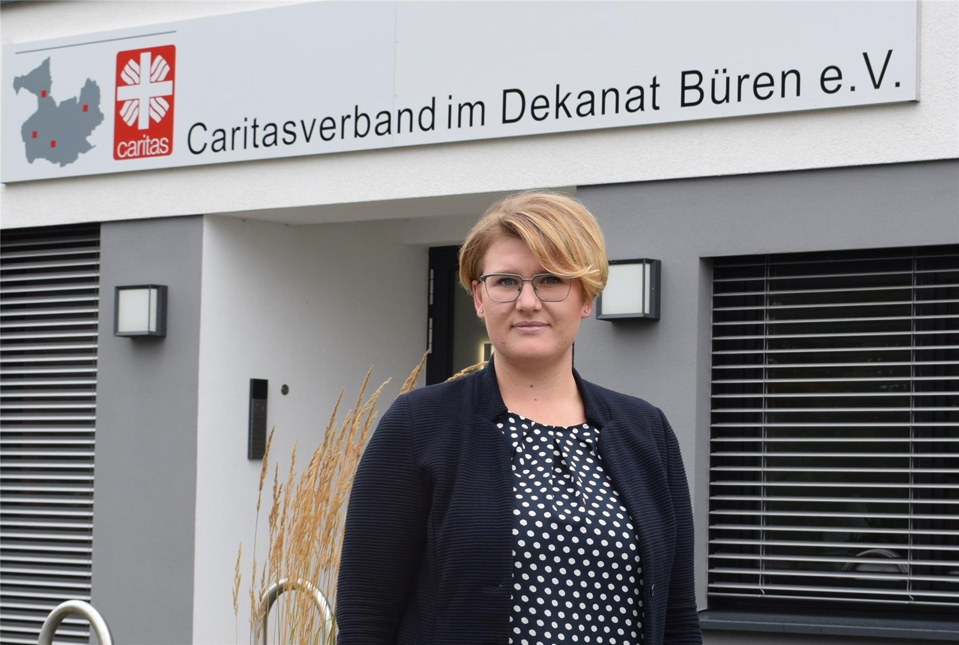 Frau mit kurzem blonden Haar, Brille und dunklem Oberteil steht vor dem Caritasgebäude