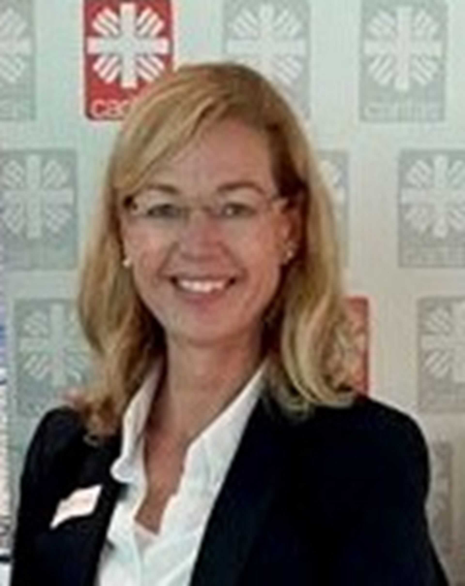 Frau mit langem blonden Haar und Brille sowie dunklem Oberteil steht vor einer Leinwand mit Caritaslogos