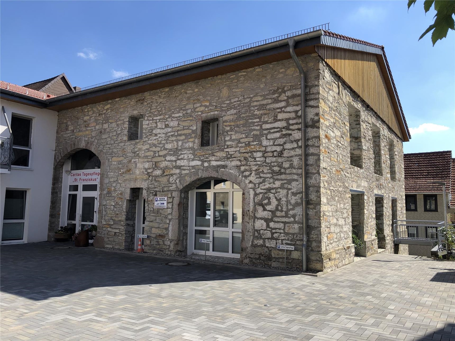 graues Bruchsteingebäude mit Holzgiebel und weiße Sprossentür und bodentiefe Fenster. Über der Tür steht in roten Buchstaben Tagespflege "St. Franziskus" Lichtenau. Hinter dem Dach des Gebäudes ist der blaue Himmel zu sehen. 