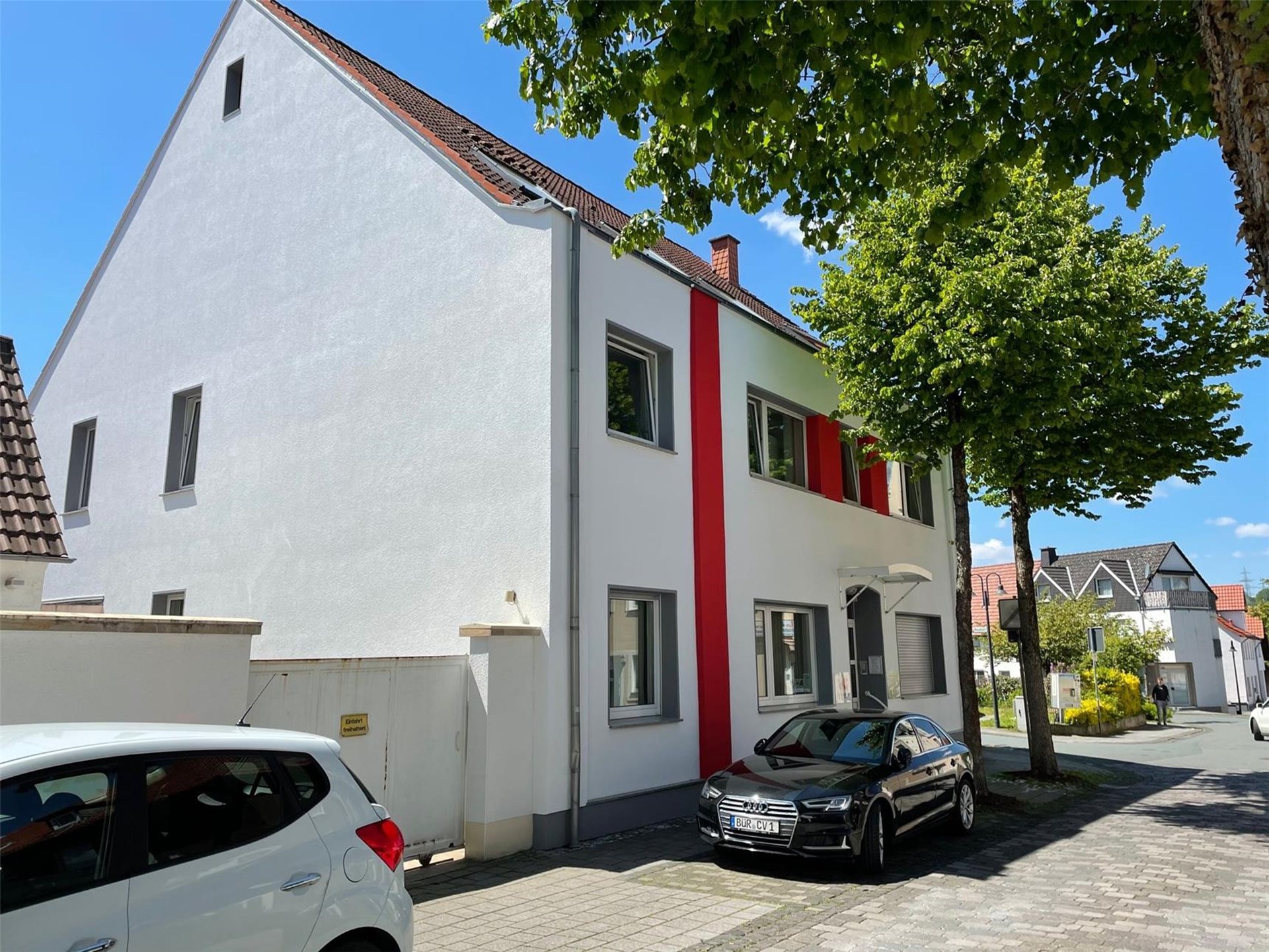 weißes Gebäude mit Spitzdach und roten Fassadenapplikationen mit grün belaubten Bäumen vor der Tür und auf dem Parkplatz stehen PKW´s