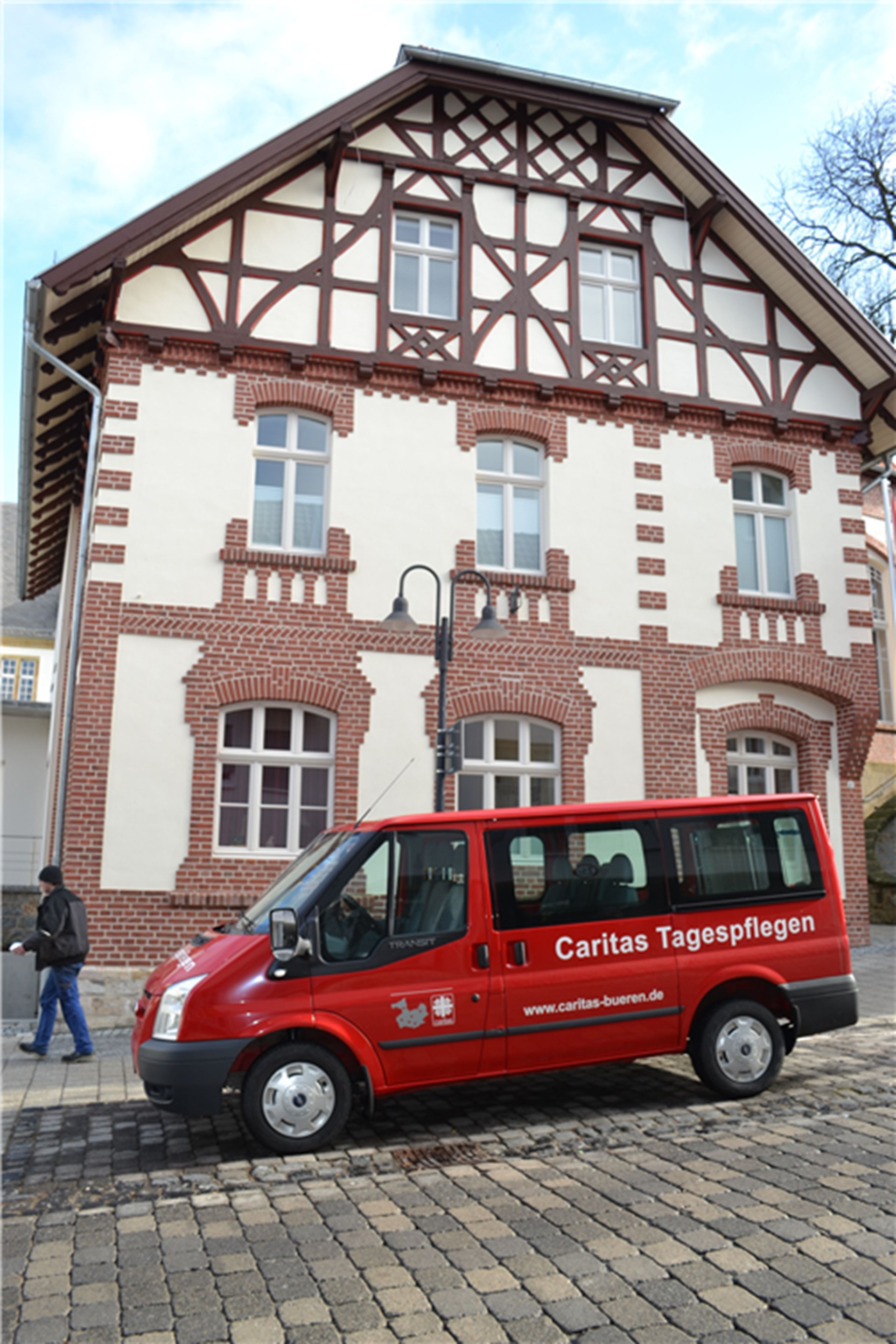 mehrstöckiges altes Gebäude mit teilweise rotem Klinker, beigefarbenden Putz und der Giebel ist aus Fachwerk. Davor steht ein rotes Behindertenfahrzeug mit weißer Aufschrift der Caritas Tagespflegen sowie das Caritas LOGO