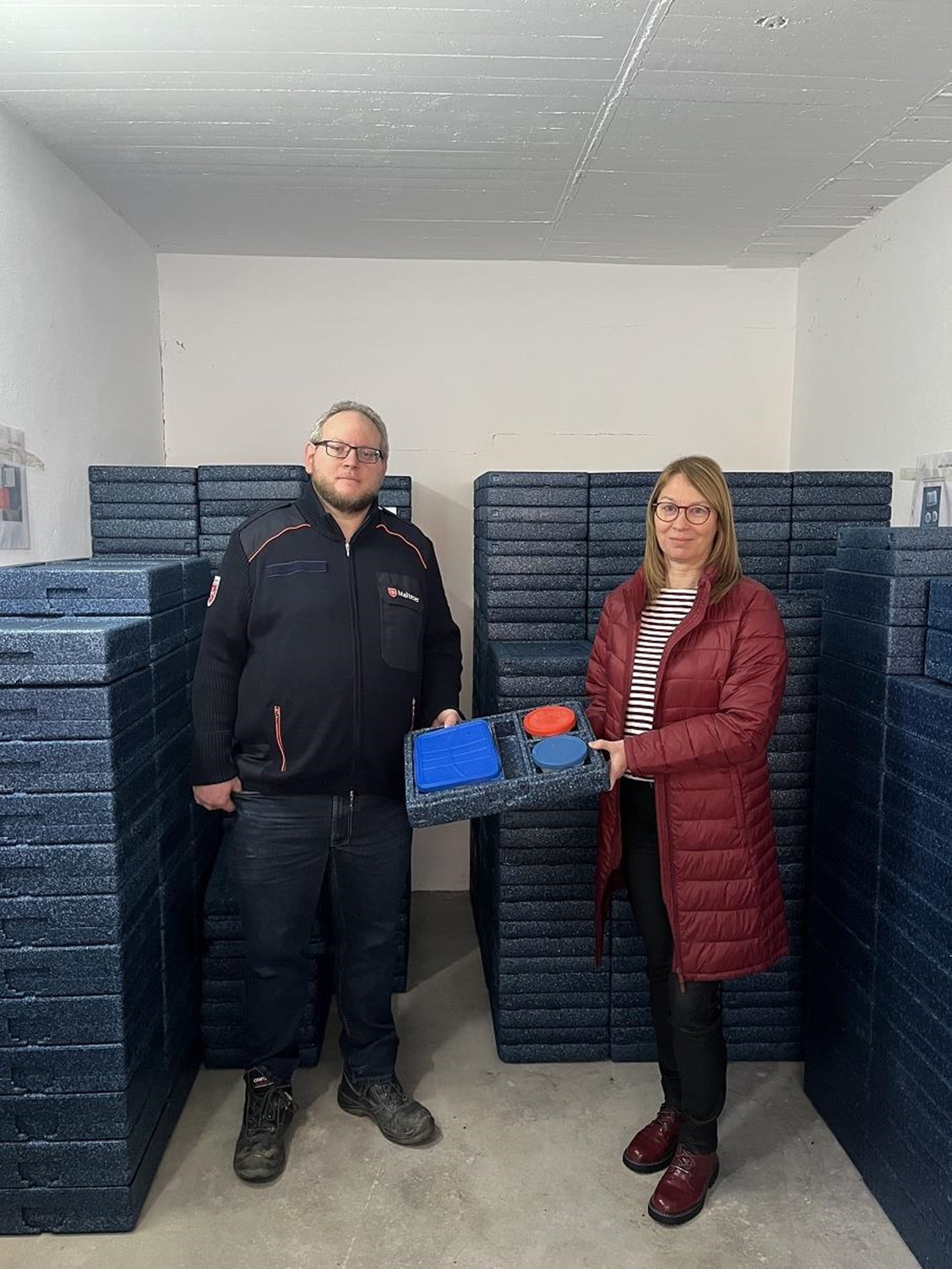 im Hintergrund des Raums stehen dunkelblaue Warmhalteboxen aus Styrophor, davor stehen ein Mann und eine Frau, die eine geöffnete blaue Box in die Mitte von sich halten