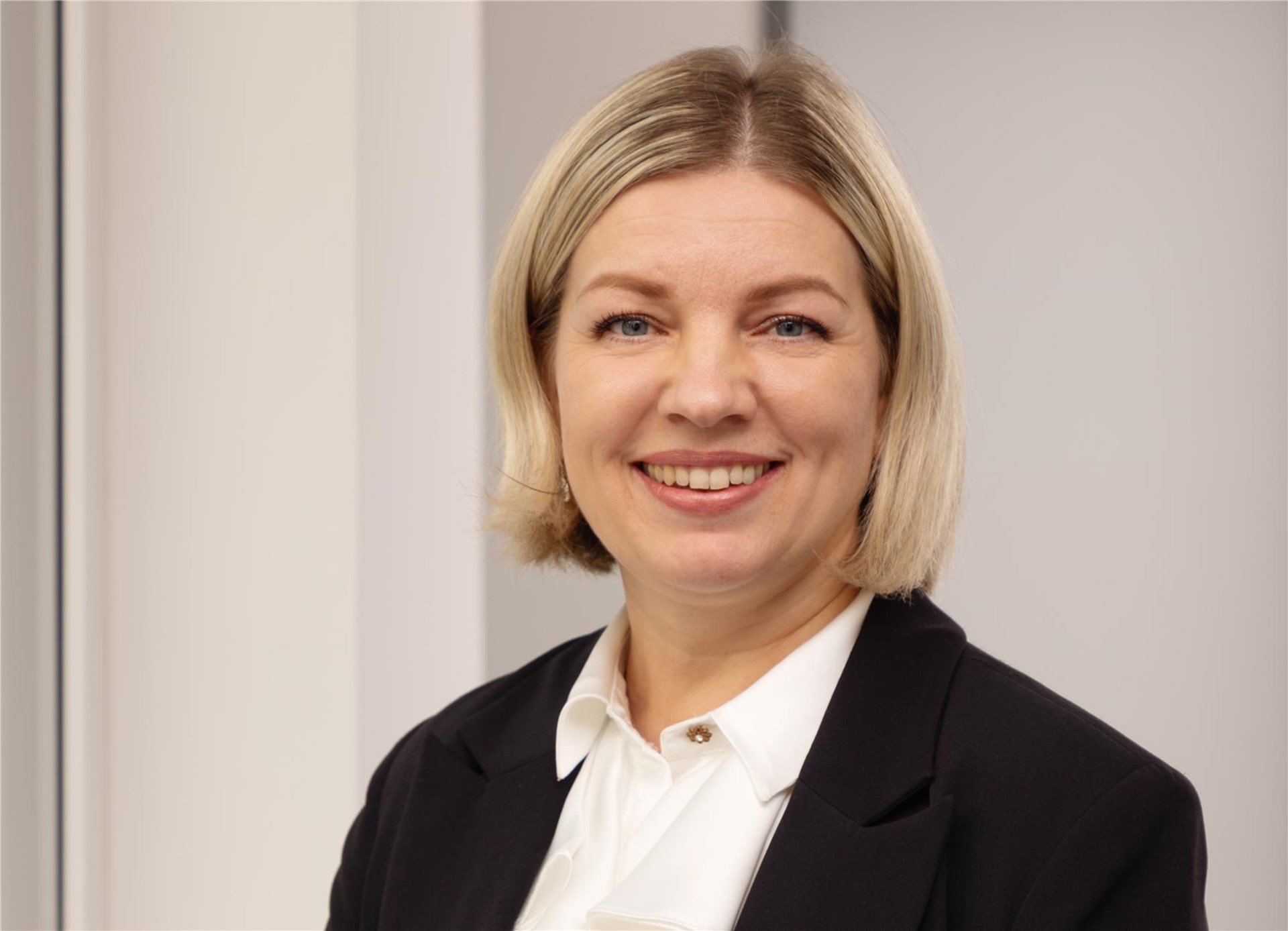 Frau mit schulterlangen blonden Haaren steht vor einem Fenster. Sie trägt eine weiße Bluse und einen schwarzen Blazer