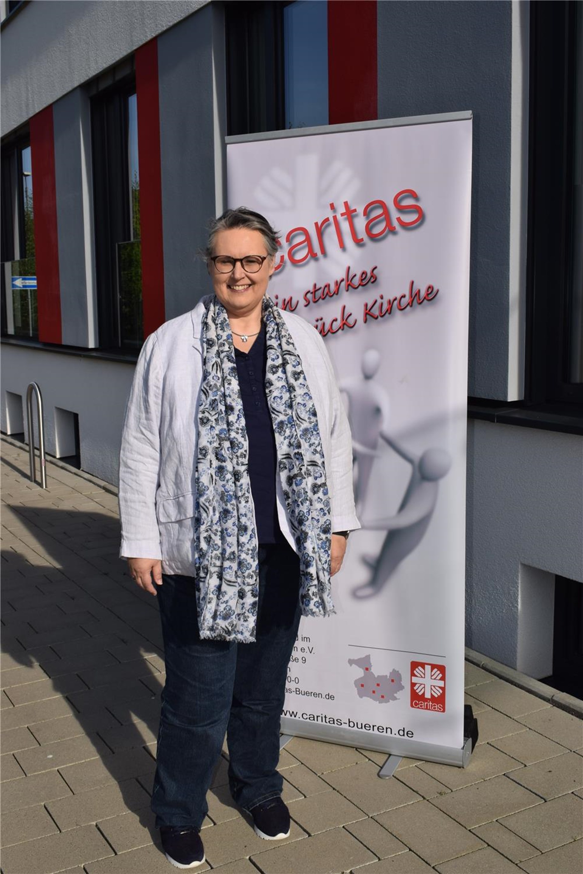 ältere Frau mit kurzem grauen Haar und Brille sowie dunkle Hose und graue längere Jacke steht vor einer Caritas Steele in weiß mit roter Schrift Ein starkes Stück Kirche
