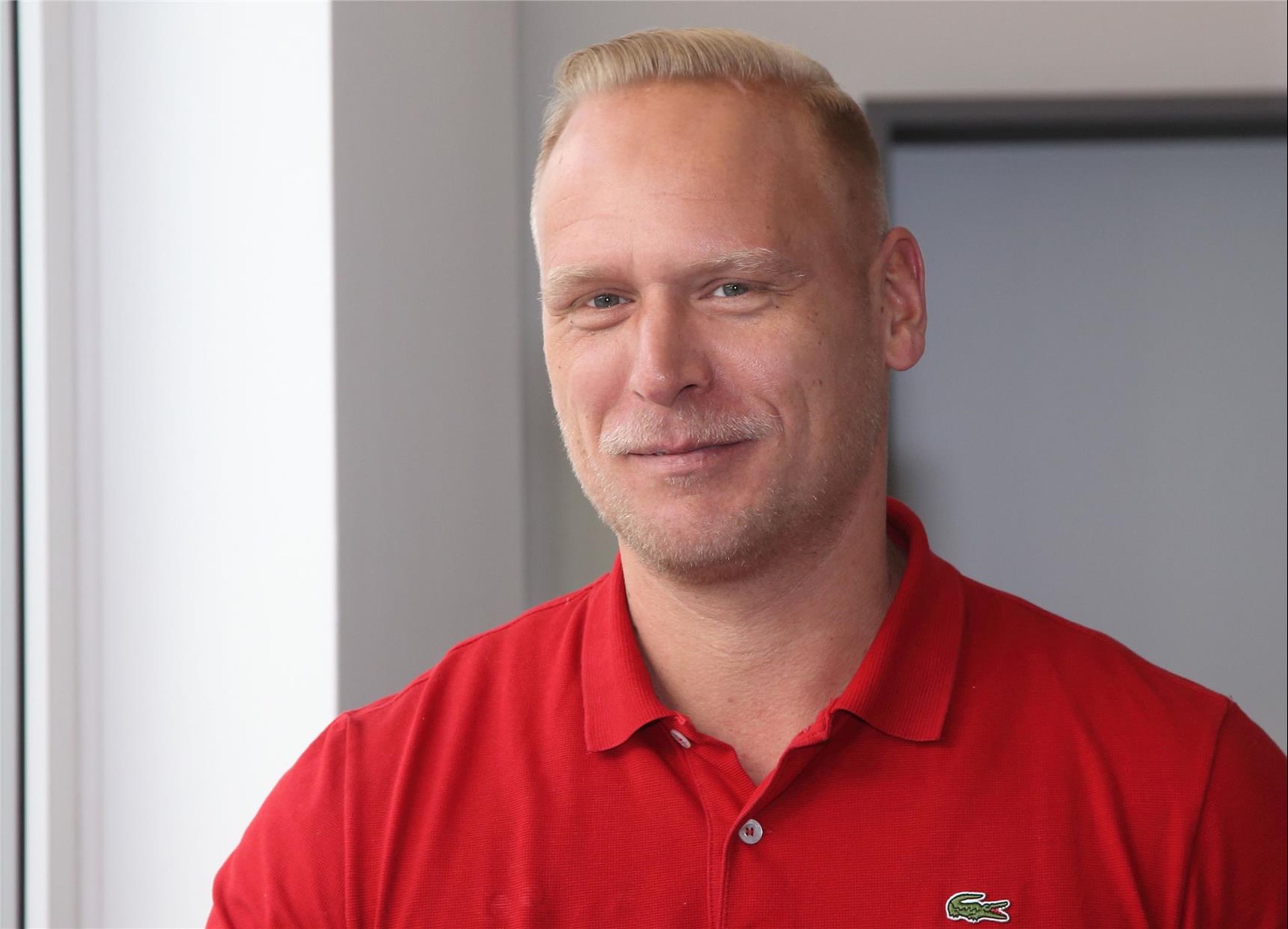 junger blonder Mann mit rotem Poloshirt steht vor einem Fenster