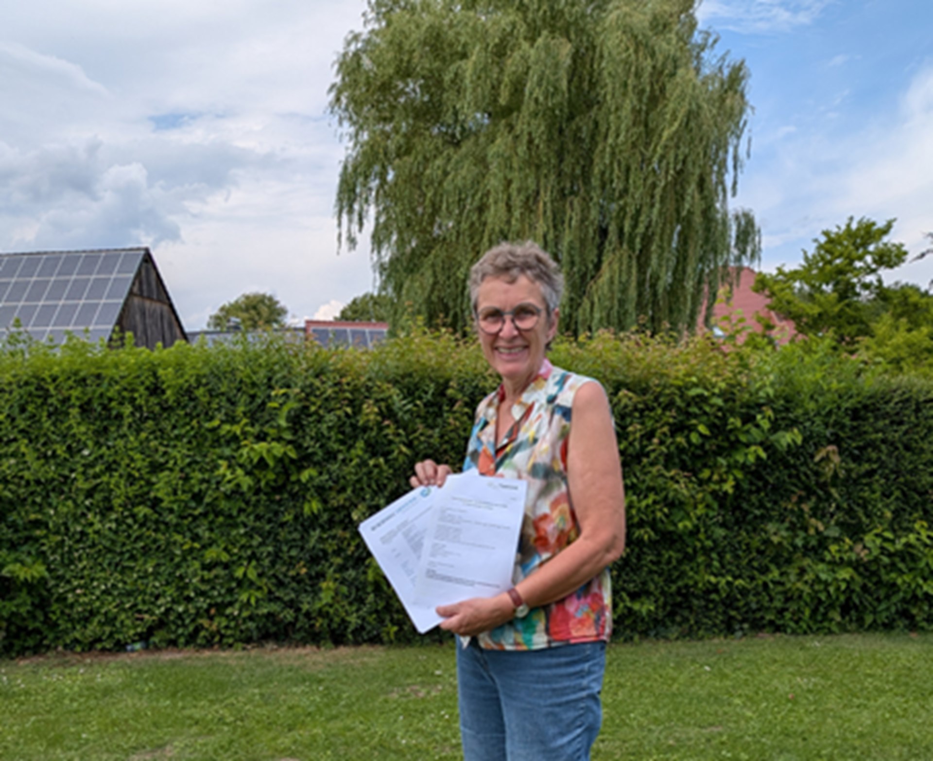 eine Frau, kurze graue Haare mit Brille, alltagstagulich gekleidet, steht auf einem Rasen und hält 2 beschriebene Papierblätter in der Hand. Vor ihr sitzt ein kleiner brauner Hund mit krausem Fell.