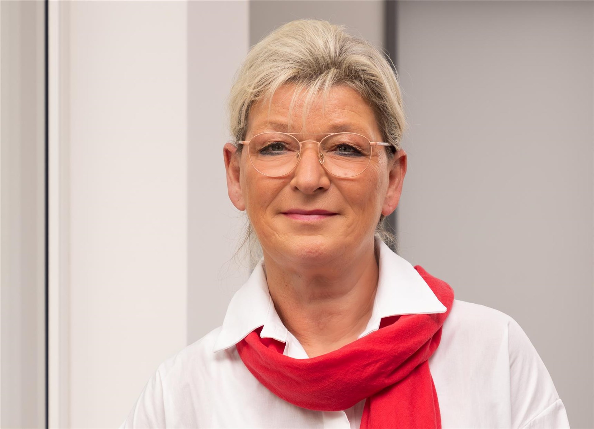 Frau mit blonden hochgesteckten Haaren steht vor einem Fenster. Sie trägt eine weiße Bluse und einen roten Schal