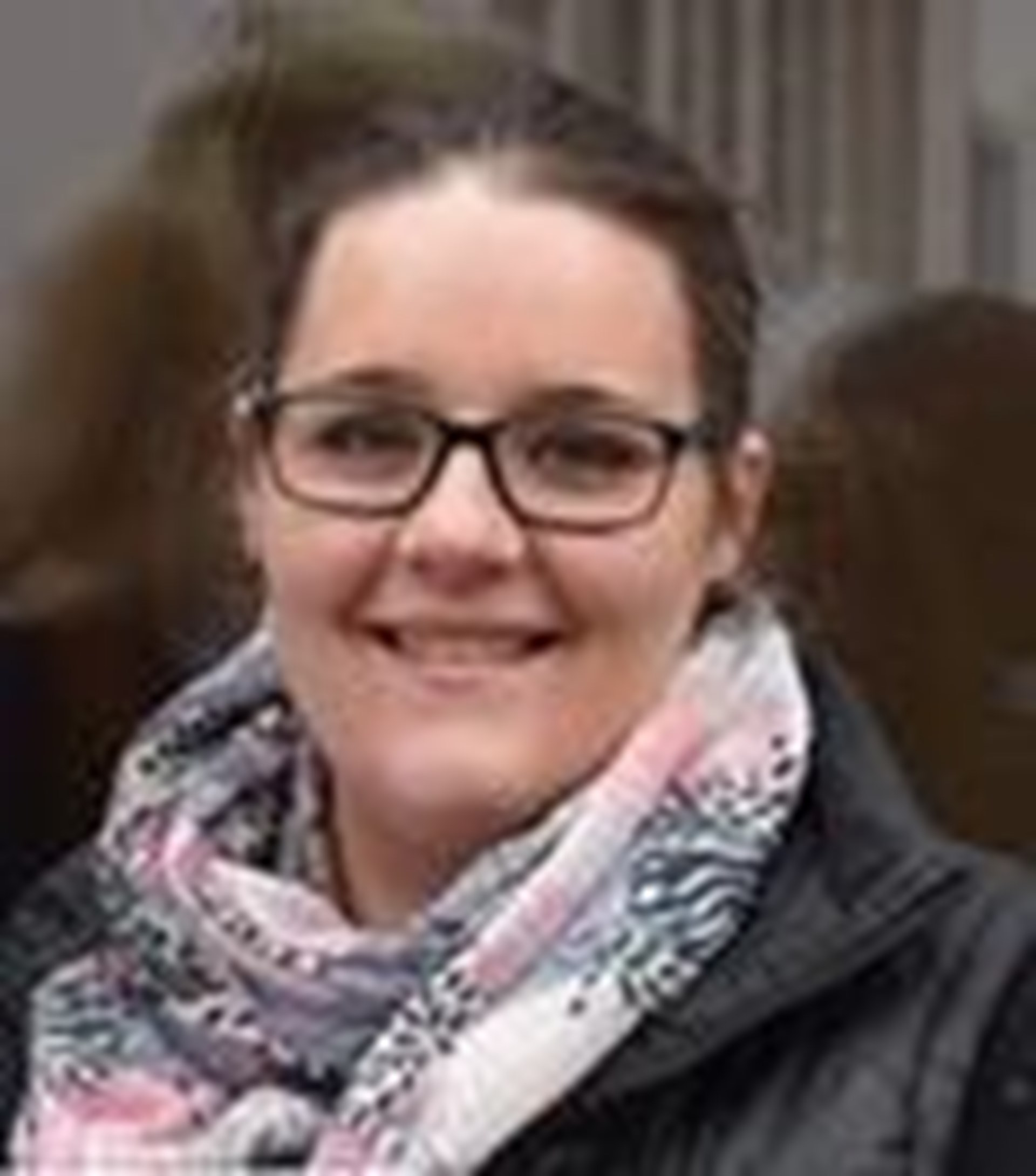junge Frau mit dunklen Haaren und Brille trägt einen bunten Schal und eine dunkle Jacke und lächelt in die Kamera