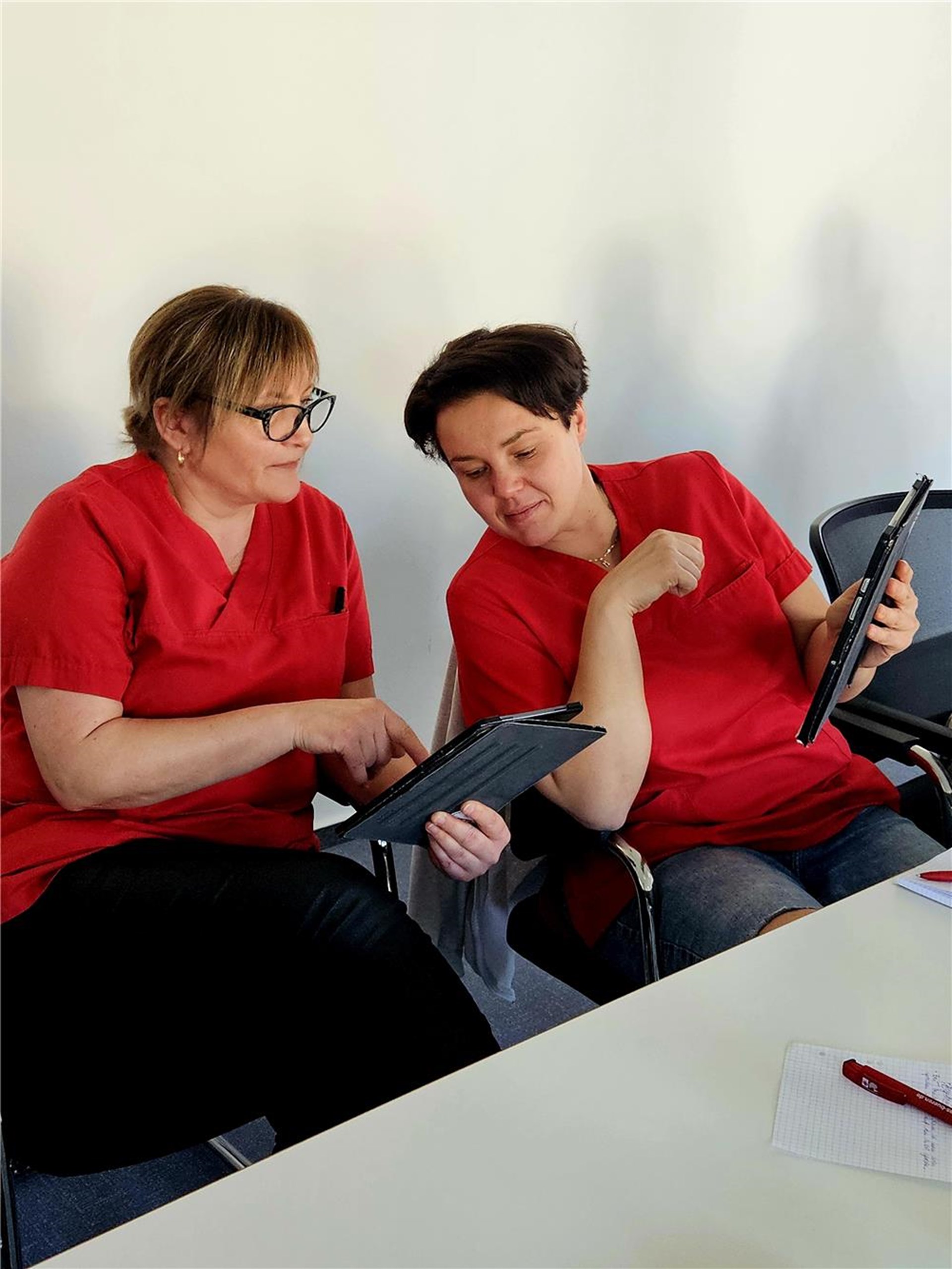 zwei Frauen im roten Oberteil sitzen an einem Tisch. beide halten ein Tablet in der Hand und schauen gemeinsam aufs Display. eine Frau hat blone Haare und trägt Brille, die andere hat dunkle kurze Haare.