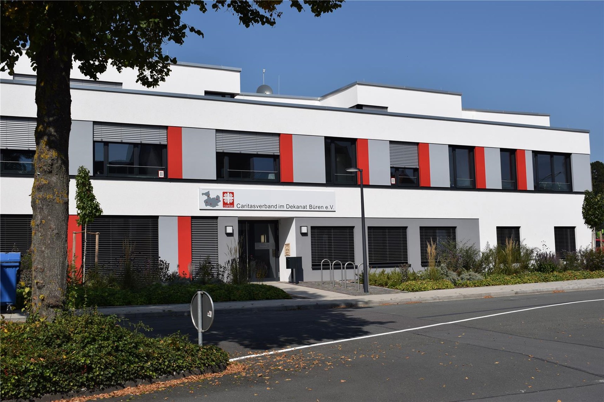 moderner Neubau mit Flachdach. Die Aussenfassade isst grau und weiß mit roten Ergänzungen. Die Eingangstür aus glas liegt etwas zurück, damit ergibt sich ein überdachter Vorsprung. Über dem Eingang befindet sich ein Schriftzug Caritasverband im Dekanat Büren e. V. 