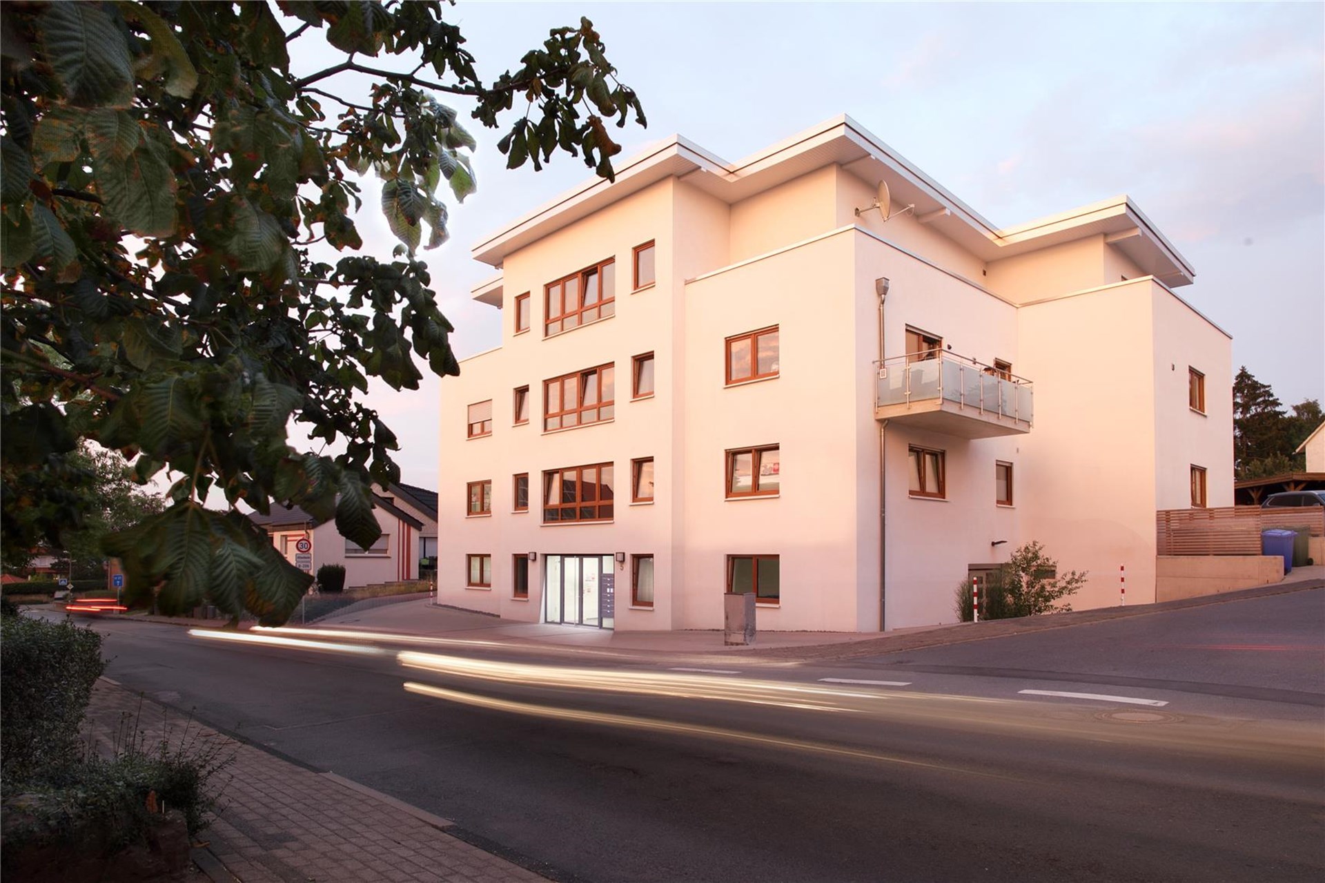 modernes Mehrparteienhaus in weiß mit Flachdach, im Vordergrund ein Baumast mit günen Blättern
