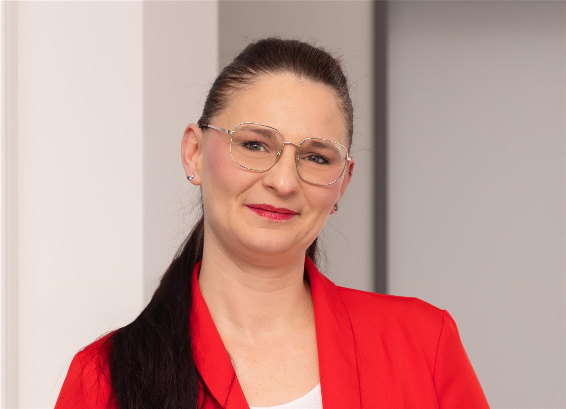 eine Frau mit Brille und dunklen, hochgesteckten Haaren steht vor einem Fenster, Sie trägt eine weiße Bluse und eine schwarze Jacke