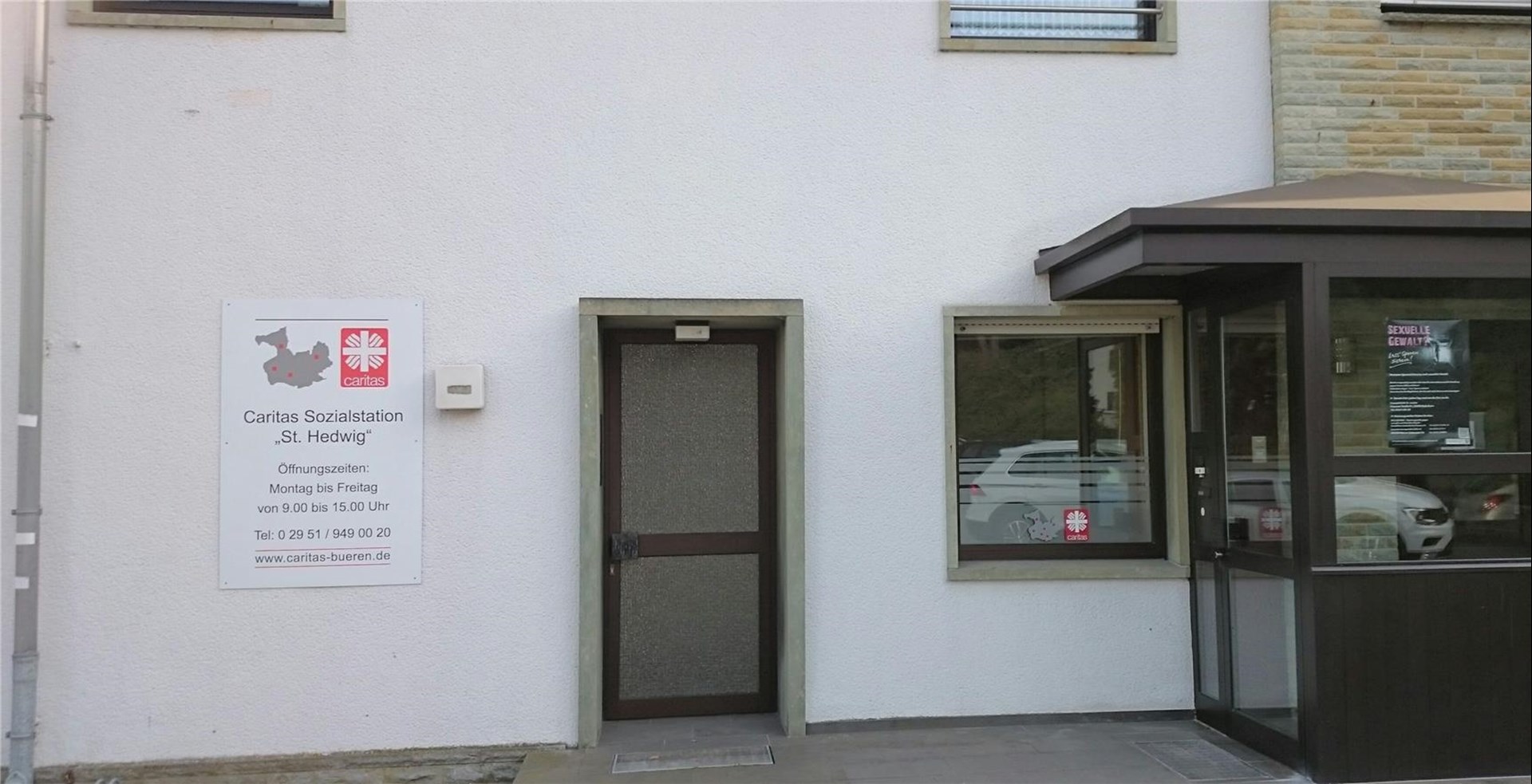 eine weiße Fassade mit einer Kontakttafel, Adresse und Telefonnummer sowie rotem Caritasverbands LOGO. Daneben eine dunkle Eingangstür mit Glas sowie einen dunkeleingefassten Windfang mit Klarglas als direkter Eingang zur Station.